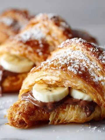 Homemade Nutella banana croissant recipe on a white marble kitchen counter, with melted chocolate and banana slices showing, dusted with powdered sugar and photographed from above.