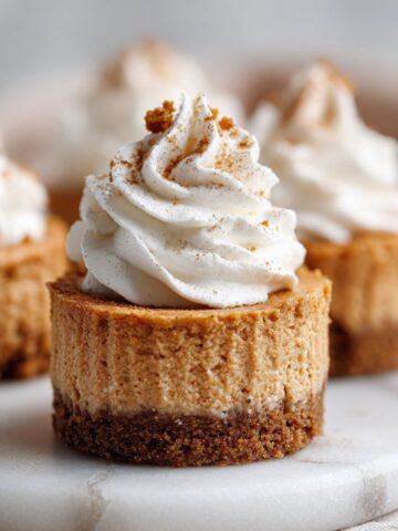 Overhead view of pumpkin spice cheesecake bites recipe ingredients on a white marble kitchen counter, including cream cheese, pumpkin puree, graham crumbs, and spices in glass bowls.