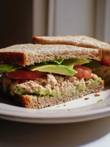 Homemade tuna avocado sandwich recipe cut in half on a plate with lettuce and tomato, taken from above on a white kitchen counter.