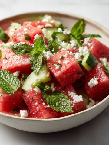 Homemade watermelon mint salad recipe with feta and cucumber served in a glass bowl on a white marble counter, photographed from above.