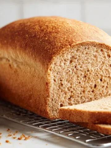 Homemade whole wheat sandwich bread recipe loaf sliced on a cooling rack with a soft fluffy texture, crumbs scattered on a white kitchen counter.