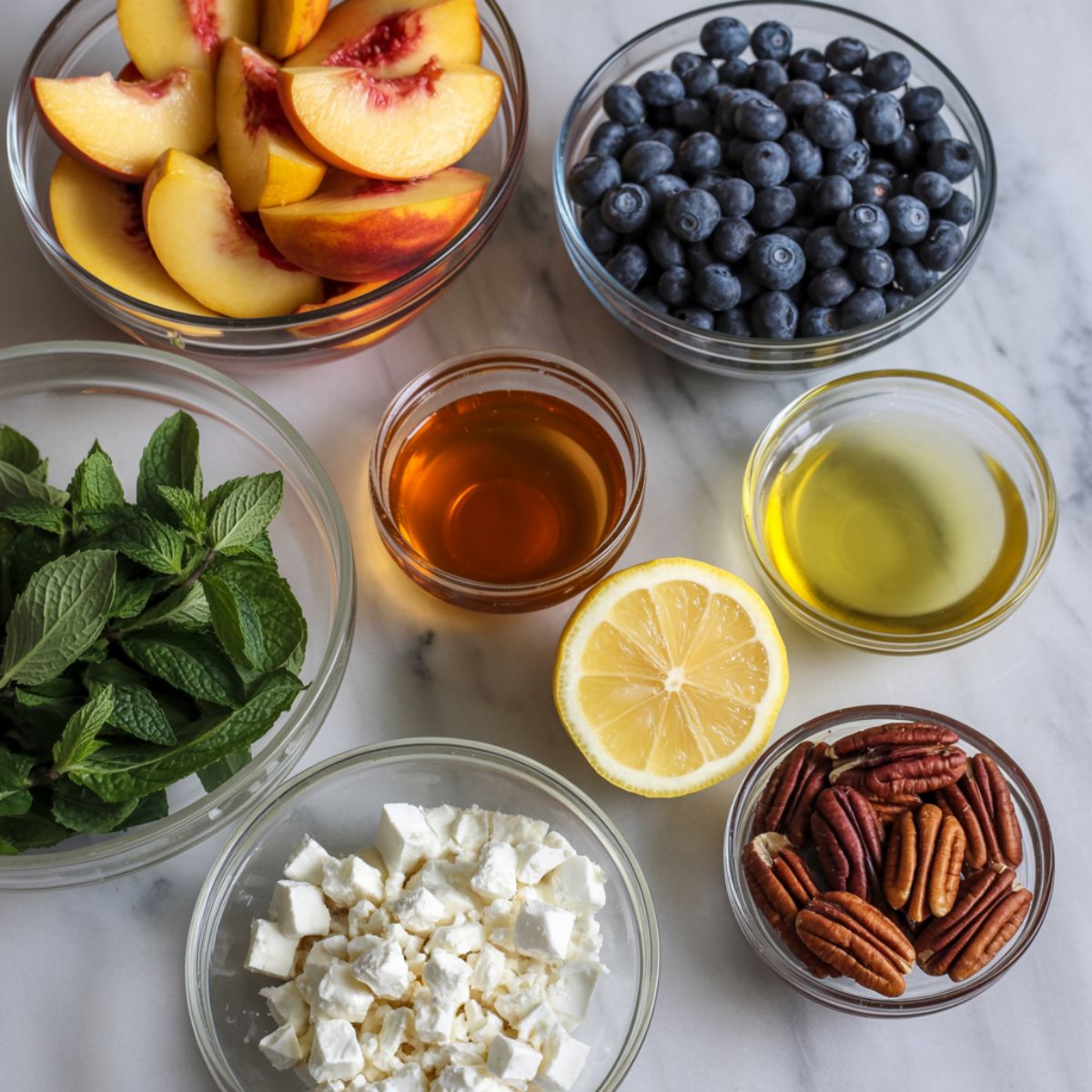 Overhead view of fresh ingredients for blueberry peach salad, including peaches, blueberries, honey, lemon, mint, feta, and pecans on a white marble kitchen counter.