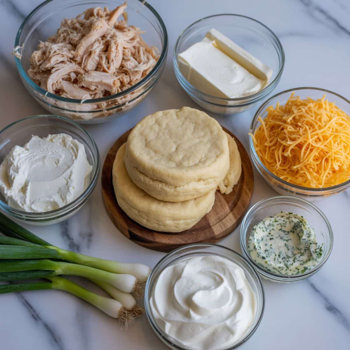 Overhead view of chicken cup ingredients — shredded chicken, cream cheese, cheddar, biscuit dough, and seasonings — on a white kitchen counter in natural light.