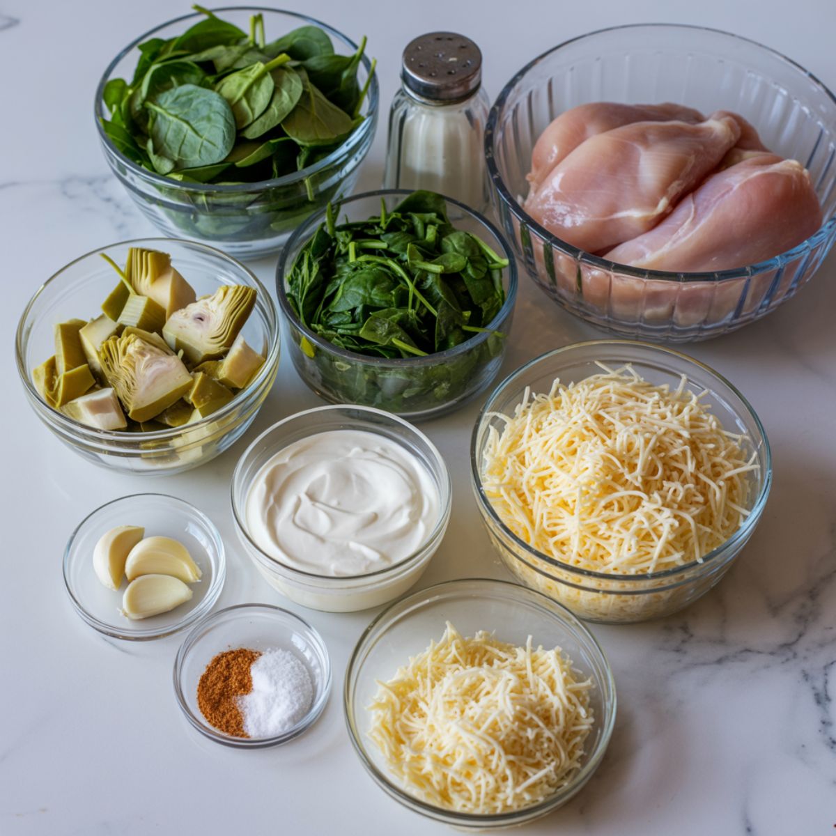 Overhead flat lay of spinach artichoke chicken casserole ingredients on a white marble kitchen counter, including chicken, spinach, artichokes, and cheese