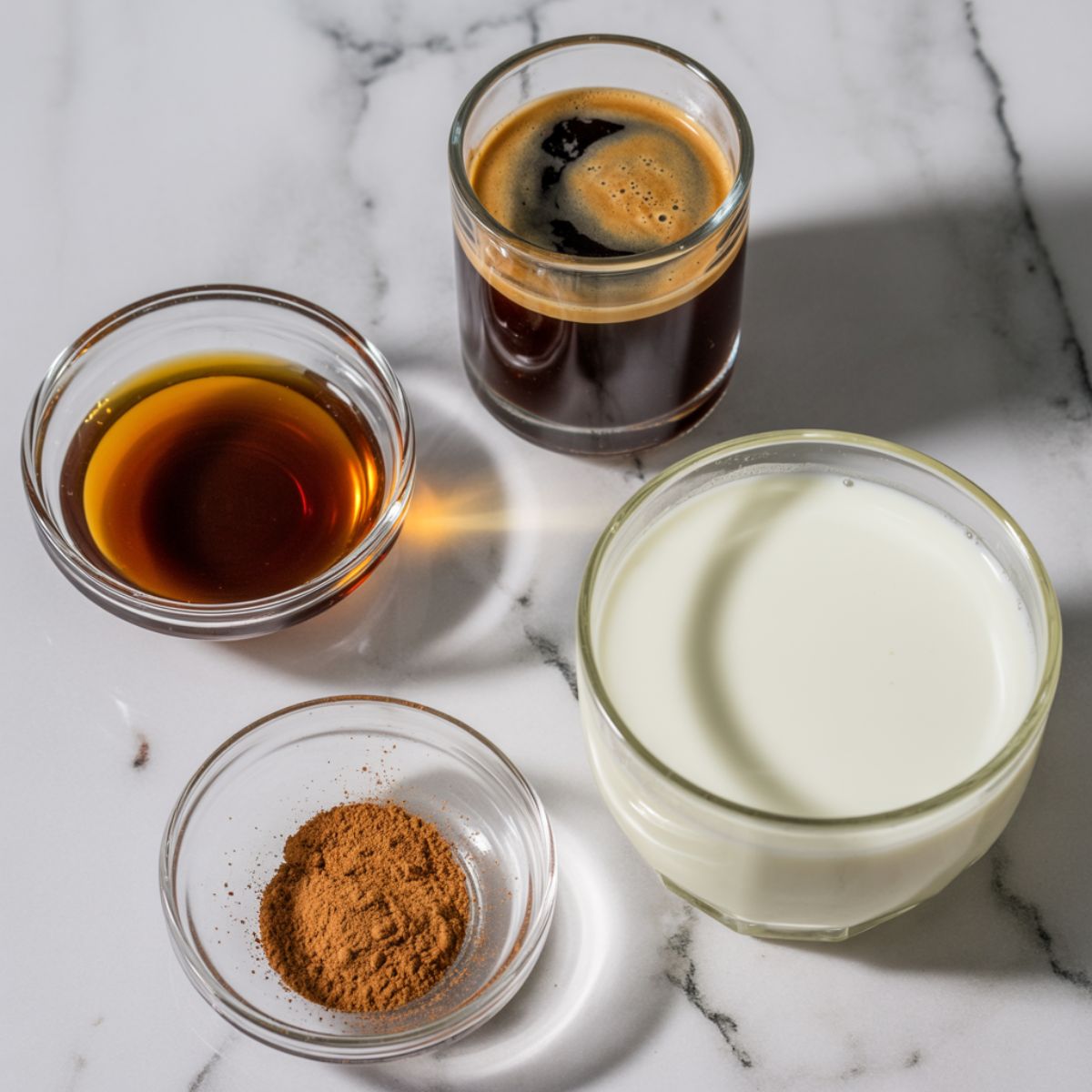 Overhead view of hazelnut cappuccino ingredients on a white marble counter including espresso, hazelnut syrup, milk, and cinnamon.