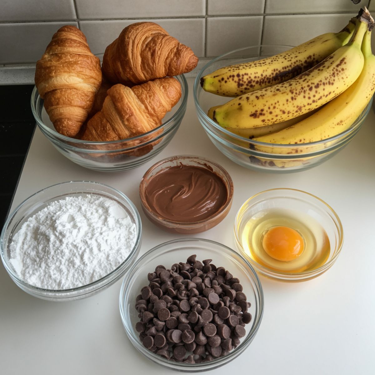 “Overhead photo of croissants, bananas, Nutella, and baking ingredients laid out on a white kitchen counter, casually arranged with a natural homemade feel.”