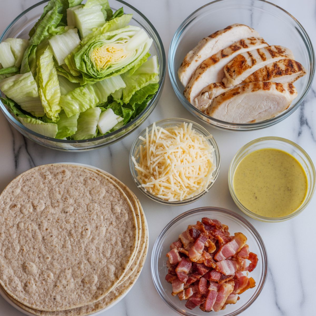 Overhead photo of chicken Caesar wrap ingredients on a white marble counter including romaine lettuce, grilled chicken, parmesan cheese, dressing, tortillas, and bacon.