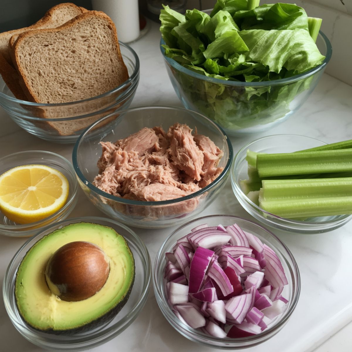 Overhead view of tuna avocado sandwich ingredients including tuna cans, avocado, lemon, onion, celery, bread, and lettuce on a white kitchen counter.