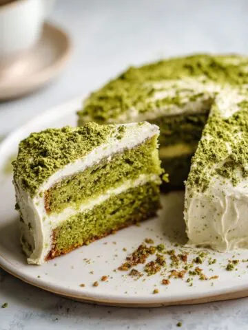 Overhead shot of a homemade matcha green tea cake recipe on a white marble counter, with a slice cut out showing its soft green crumb and slightly rustic frosting.