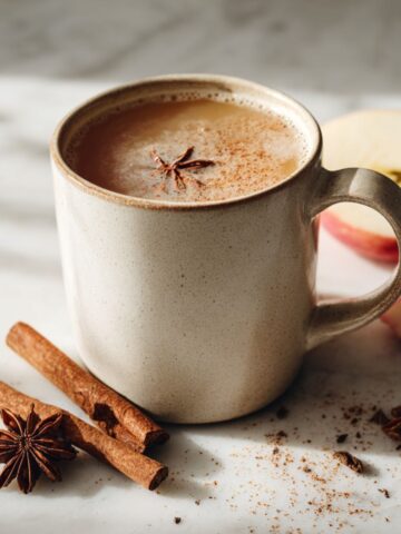 A steaming mug of spiced apple cider tea recipe on a white marble kitchen counter, surrounded by cinnamon sticks, apple slices, and cloves, photographed from above in natural morning light.