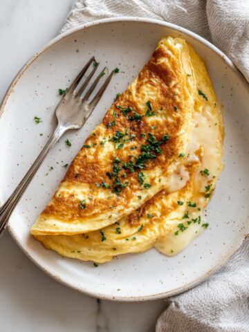 Homemade Swiss omelette recipe folded with melted Gruyère cheese on a white plate, photographed from above on a white marble counter.