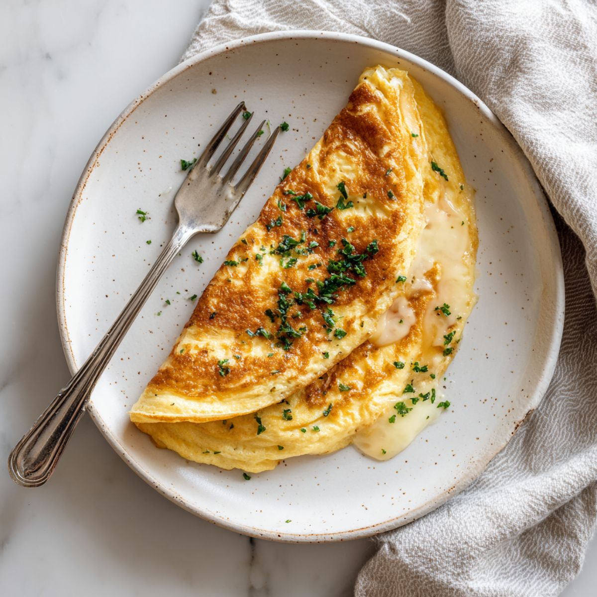Homemade Swiss omelette recipe folded with melted Gruyère cheese on a white plate, photographed from above on a white marble counter.