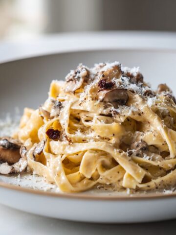 Homemade truffle tagliatelle recipe pasta served on a plate with creamy sauce, mushrooms, and Parmesan on a white kitchen counter.