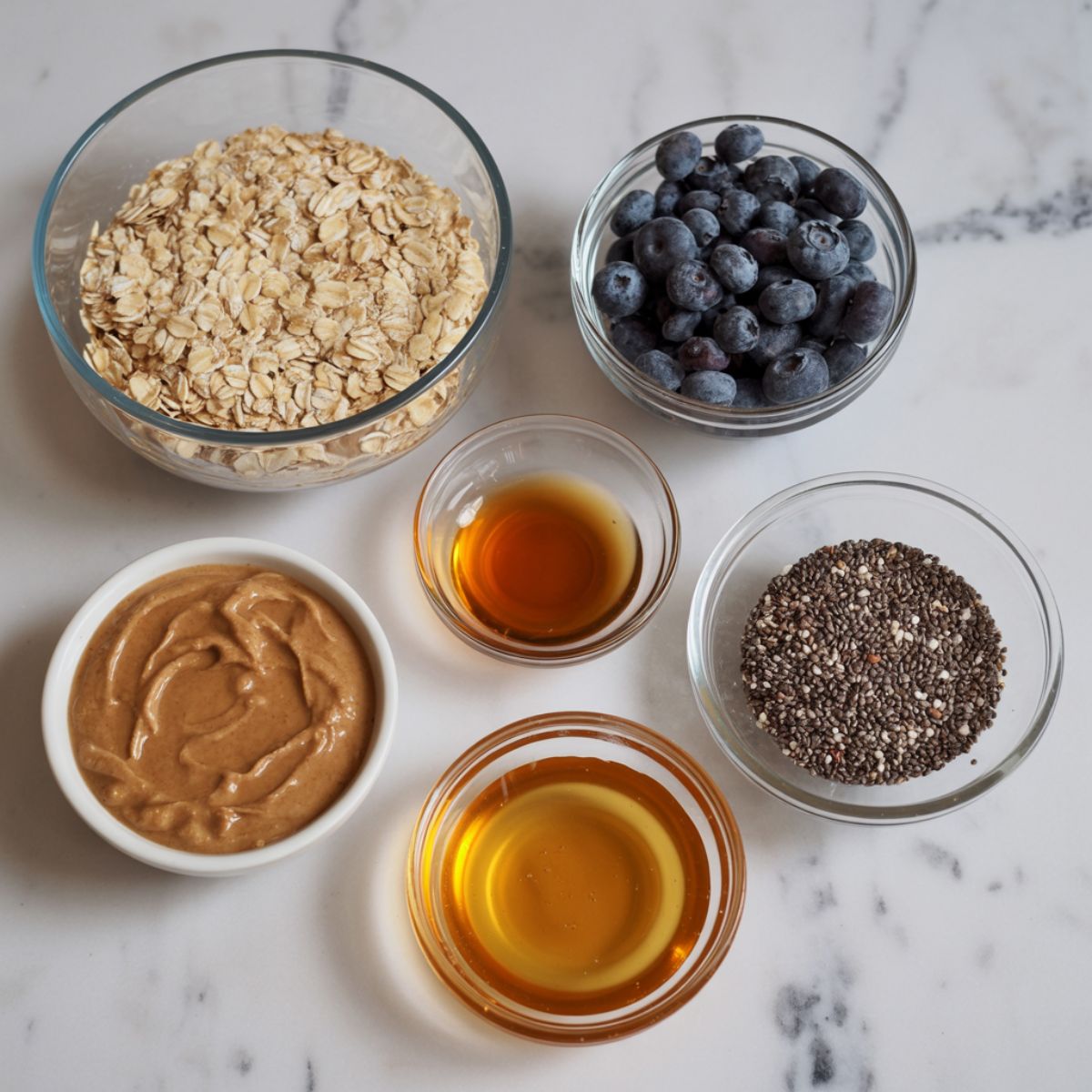 Overhead shot of ingredients for blueberry energy balls on a white marble counter, including oats, dried blueberries, almond butter, honey, and chia seeds.