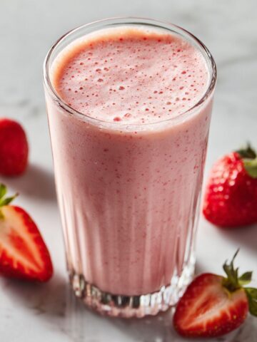 “Homemade strawberry banana protein smoothie recipe in a clear glass on a white kitchen counter with strawberries, banana slices, and protein powder around it.”