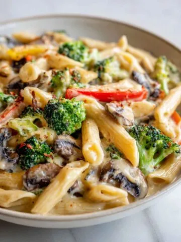 Homemade white sauce pasta with vegetables recipe served in a shallow bowl on a white marble counter, showing creamy sauce and colorful veggies from an overhead angle.