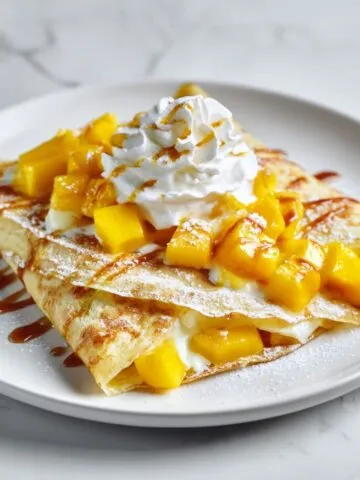 Overhead shot of a homemade mango pancake recipe filled with whipped cream and mango pieces on a white marble kitchen counter.